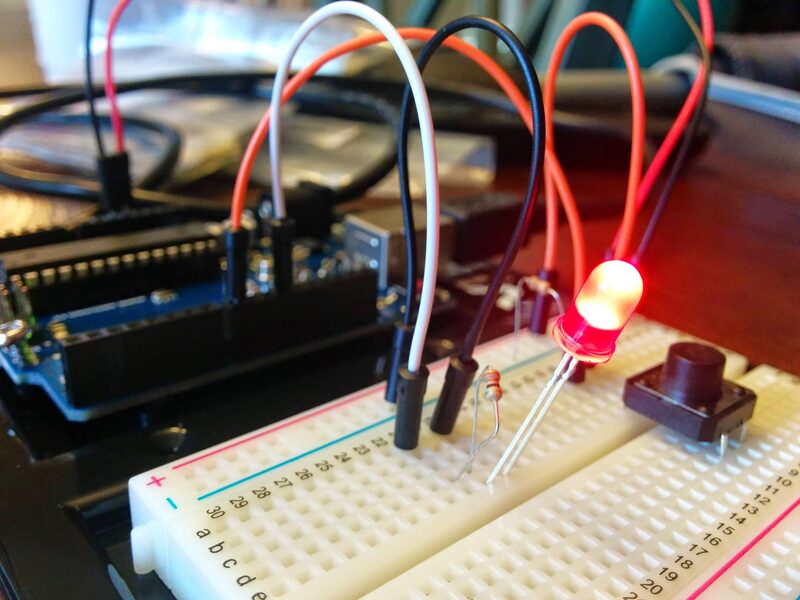 A breadboard circuit with an LED lit up red, a pushbutton, and a microcontroller board