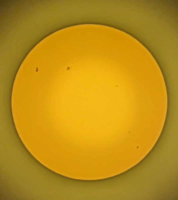 The sun through a solar filter, orange disk with small dark sunspots visible