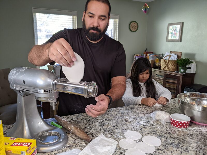 Me feeding dough through a pasta roller attachment on a stand mixer, with my daughter working dough beside me at the counter