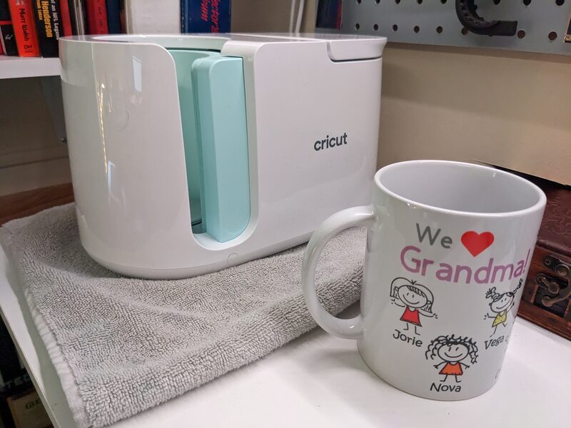 A Cricut mug press next to a finished personalized mug made as a gift for grandma