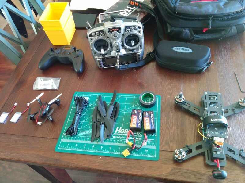 Drone parts laid out on a table: an FrSky Taranis radio, a Fatshark goggles case, a small toy quadcopter, LiPo batteries, propellers, and a partially assembled racing frame