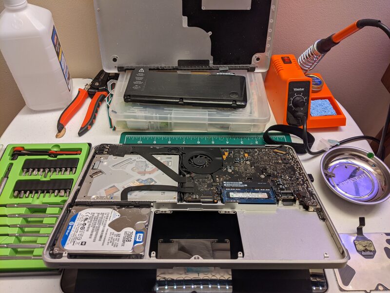 A MacBook opened up for repair - battery removed, hard drive and logic board exposed, precision bit set, a hot air rework station, and a UV curing light