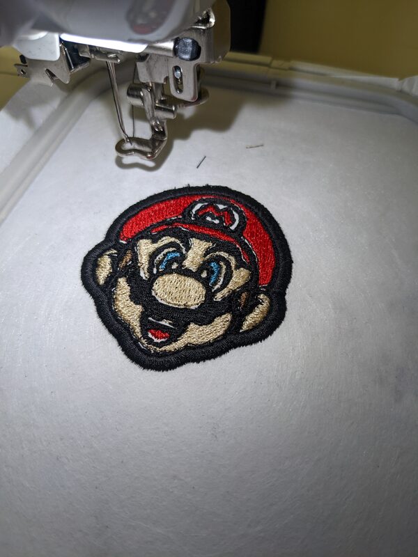 The embroidery machine mid-stitch on a black Mario face patch