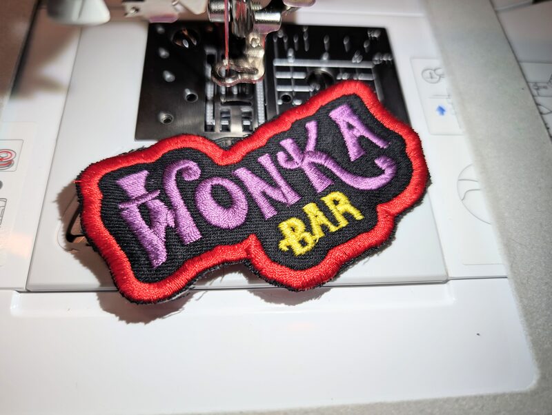 A Wonka Bar patch with purple and gold lettering on black, mid-stitch