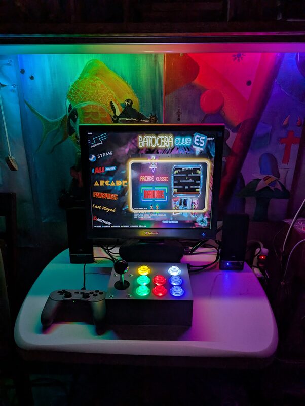 The finished Batocera setup - a white 3D printed arcade controller with color-lit buttons, RGB lighting, and framed oil paintings behind the monitor showing the Batocera menu