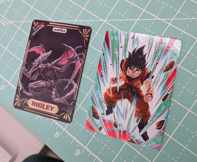 A custom holographic Goku card next to a Ridley Super Smash Bros amiibo card, on a cutting mat
