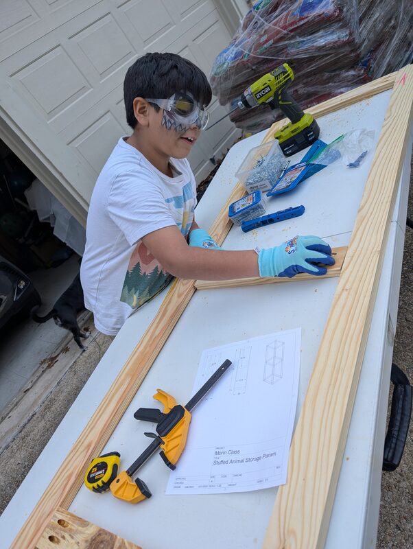 My son wearing safety glasses and running a drill, building a stuffed animal storage tower from a plan I drew up