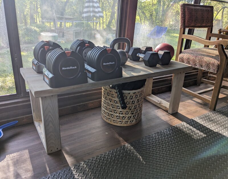 A wooden bench I built, holding adjustable dumbbells, a kettlebell, hex dumbbells, and a medicine ball in our workout room